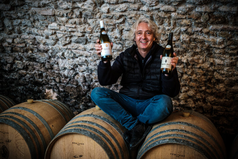 Florian Mollet sitzt auf zwei Weinfässer und hält zwei seiner Weine in den Händen